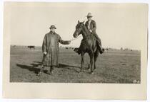 Two men, one on horseback, standing in a field with grazing cattle in the background, circa 1924  